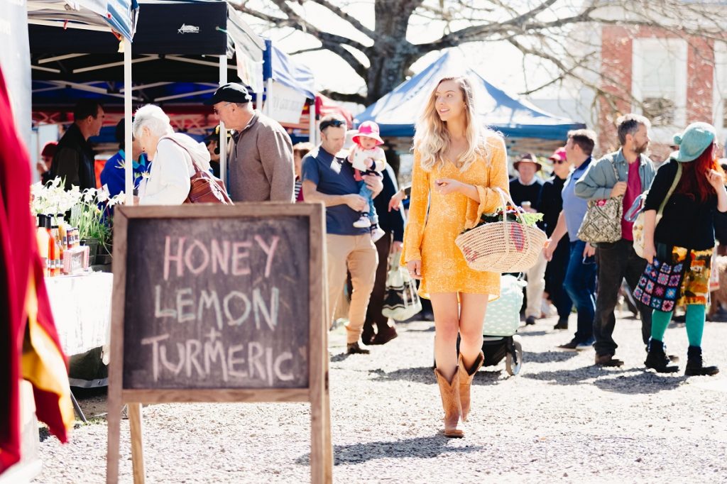 Young woman with long blond hair and long sleeve bright yellow dress with tan cowboy boots walking through Kyneton Farmers Market on an autumn day. She has a basket of vegetables hanging over one arm. People are milling around behind her and there is a stall blackboard promoting honey, lemon and tumeric. The tree behind her has lost its leaves.
